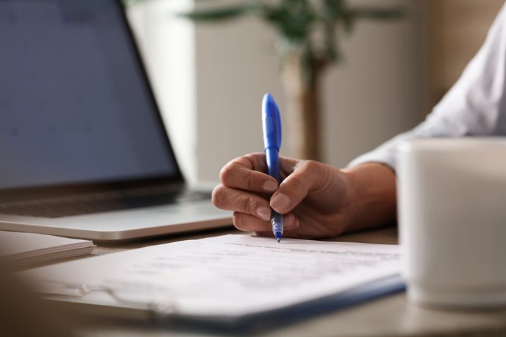 Homem escrevendo em contrato de registro de marcas na mesa de trabalho com notbook ao lado