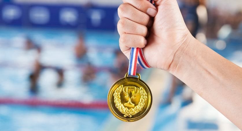 Homem segurando medalha com imagem desfocada de piscina olímpica ao fundo representando a relação entre as olimpíadas e propriedade intelectual